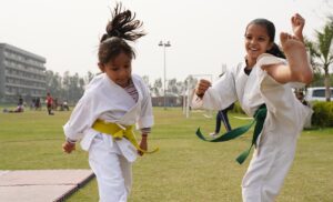 An image of two young girls in karate uniforms. One is doing a kick in the air and smiling.