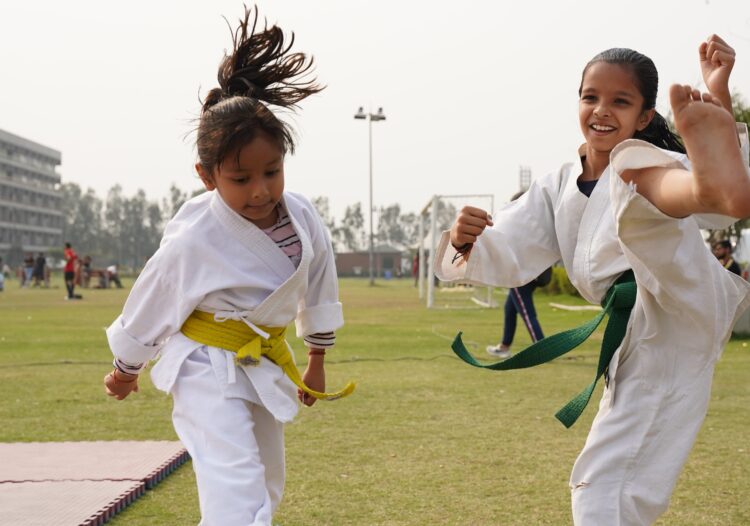 An image of two young girls in karate uniforms. One is doing a kick in the air and smiling.